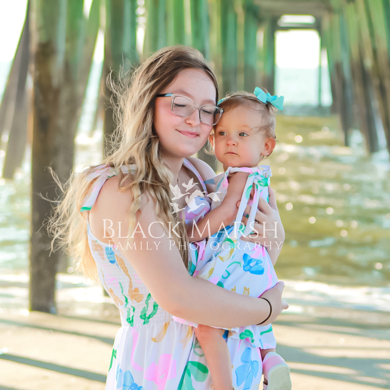 Young mom pleasantly holding her toddler daughter and posing under a pier