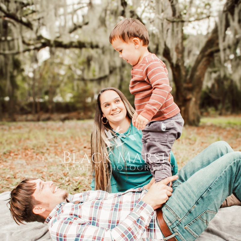Pleasant family photograph of Father holding his son up as smiling mother looks on with oak tree in background